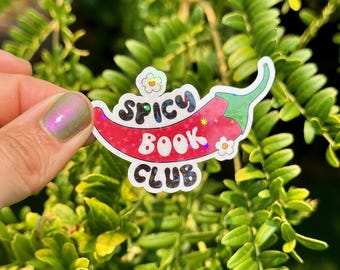 May include: A colorful sticker with the text "Spicy Book Club" in a playful font. The design features a red chili pepper with a green stem and small flower accents. The sticker is held against a backdrop of green foliage.