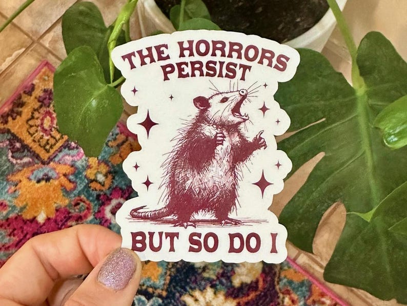May include: A white sticker with a red opossum illustration and the text "The Horrors Persist But So Do I".