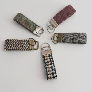 May include: Five fabric keyrings in different patterns and colours, including green, burgundy, and houndstooth. Each keyring has a metal ring for attaching keys. The keyrings are arranged on a white surface.