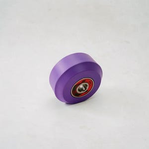 May include: A single purple roller skate wheel with a red and silver bearing. The wheel is a solid, vibrant purple color and is designed for roller skating. The bearing is visible in the center of the wheel.