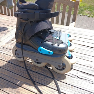 Puede incluir: Patines en línea negros con marcos grises y ruedas blancas. Los patines tienen cordones negros, correas y un acento azul. Los patines están sobre una superficie de madera.