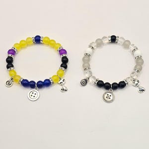 May include: Two beaded bracelets with charms. One bracelet has yellow, purple, and blue beads with a button charm and a cat charm. The other bracelet has white, black, and gray beads with a button charm and a cat charm.