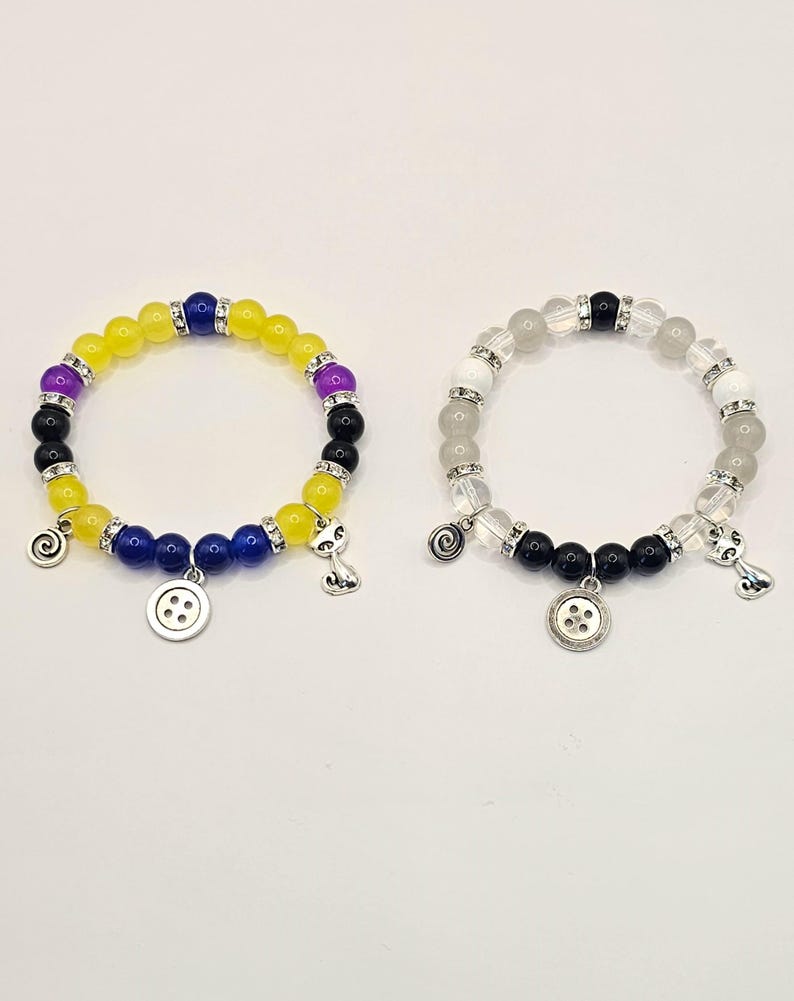 May include: Two beaded bracelets with charms. One bracelet has yellow, purple, and blue beads with a button charm and a cat charm. The other bracelet has white, black, and gray beads with a button charm and a cat charm.