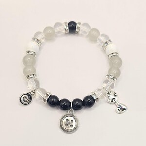 May include: A white and black beaded bracelet with a button charm, a cat charm, and a swirl charm. The bracelet is made with clear and opaque white beads, black beads, and silver accents.