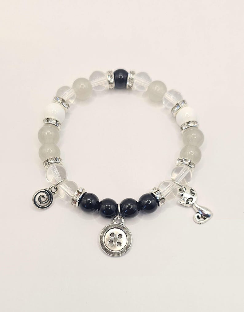 May include: A white and black beaded bracelet with a button charm, a cat charm, and a swirl charm. The bracelet is made with clear and opaque white beads, black beads, and silver accents.