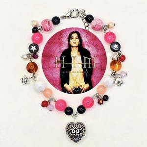 May include: A beaded bracelet featuring a central circular image of a person with the band name "HIM" and album title "Razorblade Romance". The bracelet includes pink, black, and white beads, star charms, and a silver heart pendant.