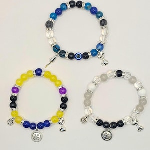 May include: Three beaded bracelets with different colored beads and charms. The first bracelet has blue, black, and clear beads with a blue evil eye charm. The second bracelet has yellow, purple, blue, and black beads with a button charm. The third bracelet has white, black, and clear beads with a swirl charm.