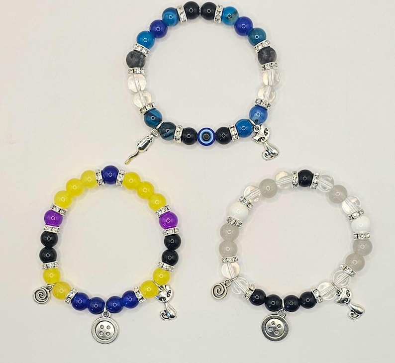 May include: Three beaded bracelets with different colored beads and charms. The first bracelet has blue, black, and clear beads with a blue evil eye charm. The second bracelet has yellow, purple, blue, and black beads with a button charm. The third bracelet has white, black, and clear beads with a swirl charm.