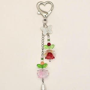 May include: A silver heart-shaped keychain with a silver butterfly charm and two layered flower charms. The flowers are made of pink, red, and green glass beads.