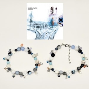May include: Two beaded bracelets with a variety of beads in shades of blue, gray, white, and black. The bracelets have star-shaped charms and are displayed with the album cover for Radiohead's "OK Computer."