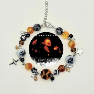 May include: A beaded bracelet featuring a mix of round and irregular beads in shades of orange, brown, black, and white. The bracelet includes star-shaped charms and a circular pendant with the text "SOUNDGARDE".