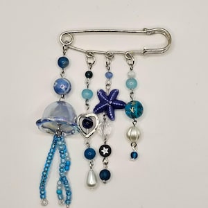 May include: A decorative brooch featuring a silver safety pin base with dangling charms. The charms include blue beads, a jellyfish, a starfish, and a heart. The color palette consists of blues, whites, and silver, creating a nautical theme.