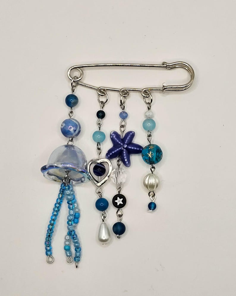May include: A decorative brooch featuring a silver safety pin base with dangling charms. The charms include blue beads, a jellyfish, a starfish, and a heart. The color palette consists of blues, whites, and silver, creating a nautical theme.