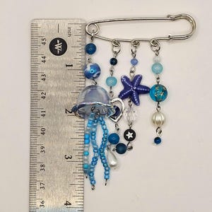 May include: A decorative pin featuring a jellyfish design with blue beads and a starfish charm. The pin is approximately 4 inches long, with a silver-tone safety pin clasp. The design includes various shades of blue and white beads.
