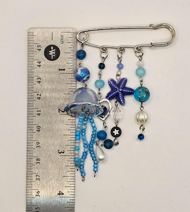 May include: A decorative pin featuring a jellyfish design with blue beads and a starfish charm. The pin is approximately 4 inches long, with a silver-tone safety pin clasp. The design includes various shades of blue and white beads.