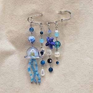 May include: A decorative brooch featuring a large silver safety pin with dangling beaded charms. The charms include a jellyfish, a starfish, and various blue beads. The brooch is set against a neutral fabric background.