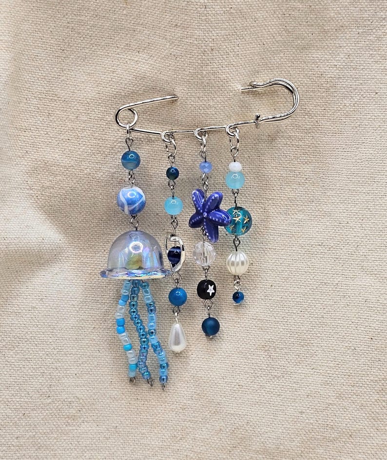 May include: A decorative brooch featuring a large silver safety pin with dangling beaded charms. The charms include a jellyfish, a starfish, and various blue beads. The brooch is set against a neutral fabric background.