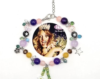 May include: A beaded charm bracelet with a central circular image of a woman wearing a crown. The bracelet features various colorful beads, including purple, pink, and yellow, along with star and flower charms. The text "Ho Live Through Th" is visible.