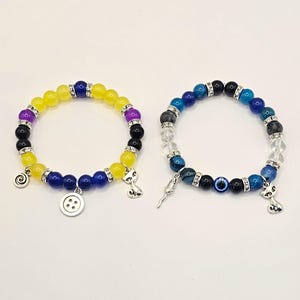 May include: Two beaded bracelets with charms. The first bracelet has yellow, purple, blue, and black beads with a button, a spiral, and a cat charm. The second bracelet has blue, black, clear, and white beads with an evil eye, a key, and a cat charm.
