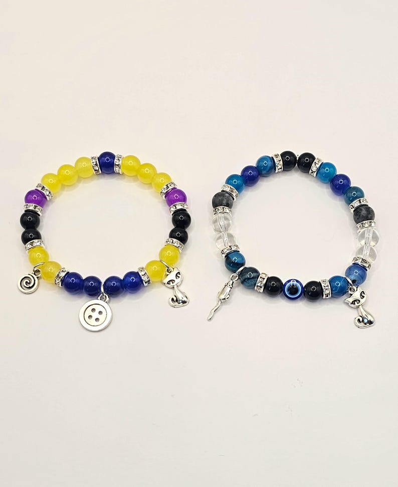 May include: Two beaded bracelets with charms. The first bracelet has yellow, purple, blue, and black beads with a button, a spiral, and a cat charm. The second bracelet has blue, black, clear, and white beads with an evil eye, a key, and a cat charm.
