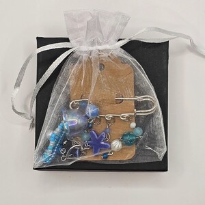 May include: A collection of blue and silver beaded jewelry, including a safety pin brooch, is presented in a sheer white gift bag. The bag is tied with a silver ribbon and placed on a small black box. The jewelry features a starfish charm.