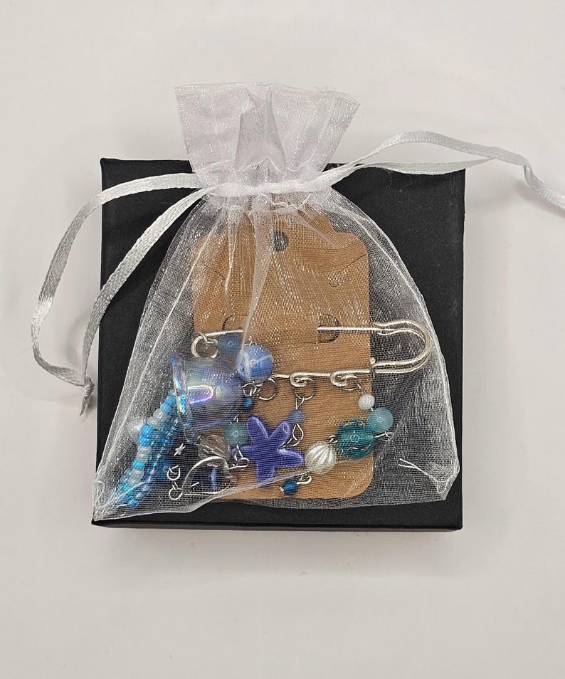 May include: A collection of blue and silver beaded jewelry, including a safety pin brooch, is presented in a sheer white gift bag. The bag is tied with a silver ribbon and placed on a small black box. The jewelry features a starfish charm.