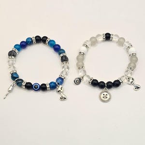 May include: Two beaded bracelets with charms. The first bracelet has blue, black, and white beads with a charm of an evil eye and a cat. The second bracelet has white beads with a charm of a button and a spiral.
