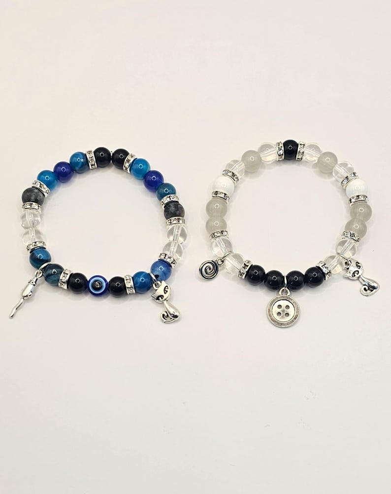 May include: Two beaded bracelets with charms. The first bracelet has blue, black, and white beads with a charm of an evil eye and a cat. The second bracelet has white beads with a charm of a button and a spiral.