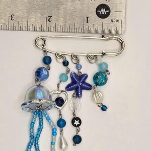 May include: A silver safety pin with dangling charms. The charms include a jellyfish, a starfish, beads, and a heart. The beads are in shades of blue, white, and clear. The ruler shows the pin is approximately 2 inches long.