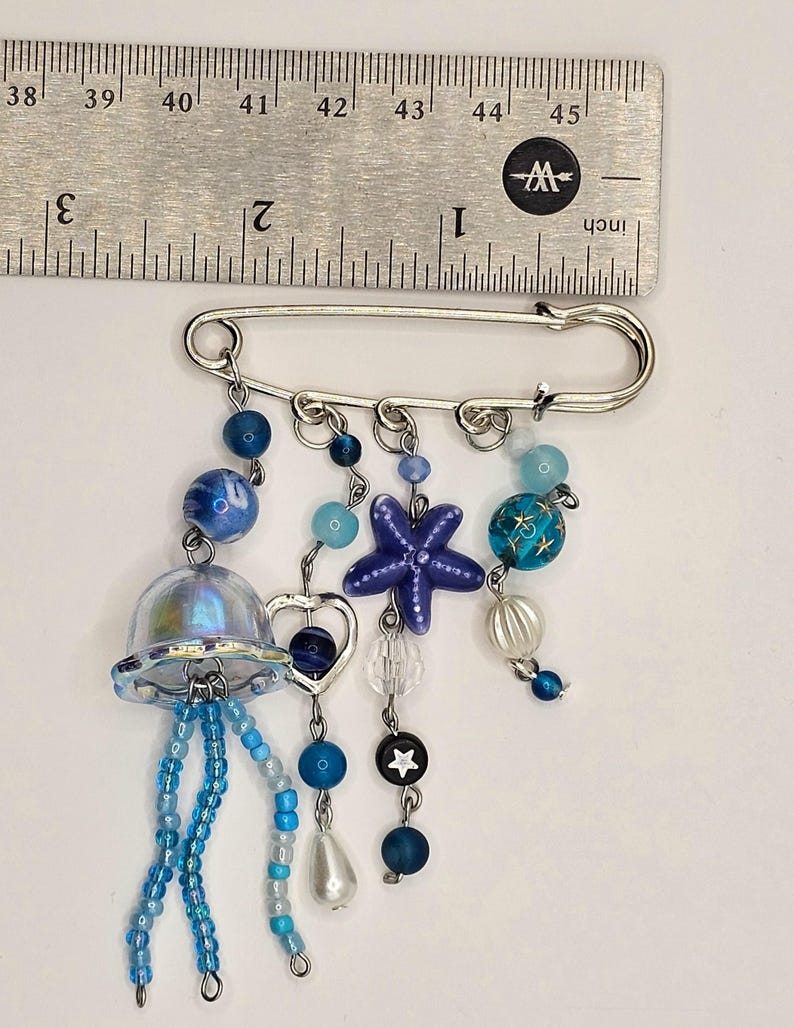 May include: A silver safety pin with dangling charms. The charms include a jellyfish, a starfish, beads, and a heart. The beads are in shades of blue, white, and clear. The ruler shows the pin is approximately 2 inches long.
