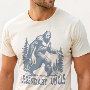 May include: Cream-colored t-shirt featuring a gray illustration of Bigfoot walking through a forest. The text "LEGENDARY UNCLE" is printed below the image in a matching gray color. The shirt is made of a soft material.