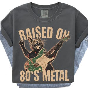 May include: Dark gray t-shirt with a graphic of a cat playing a green guitar. The text "RAISED ON 80'S METAL" is printed above and below the cat illustration. The shirt is folded over a pair of blue jeans.