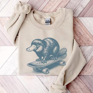 May include: A beige sweatshirt with a blue illustration of a platypus riding a skateboard. The platypus is facing left and has a long, thin tail. The skateboard has wheels and is on a light wood background.