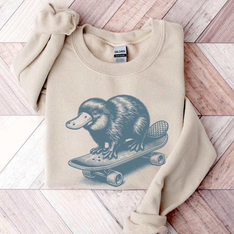 May include: A beige sweatshirt with a blue illustration of a platypus riding a skateboard. The platypus is facing left and has a long, thin tail. The skateboard has wheels and is on a light wood background.