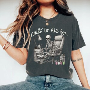 May include: Dark gray t-shirt with the phrase "nails to die for" in cursive. The graphic depicts a skeleton getting a manicure. The skeleton is seated in a salon chair, with nail polish bottles and a lamp on a table.