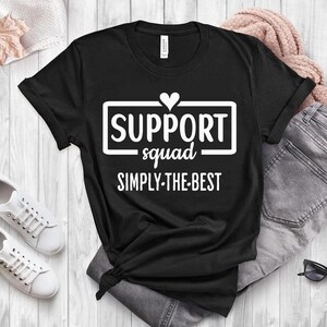May include: Black t-shirt with the text "SUPPORT squad SIMPLY-THE-BEST" in a white rectangular box with a heart above the word "SUPPORT". The shirt is styled with a pair of white sneakers, blue jeans, and a pink sweater.
