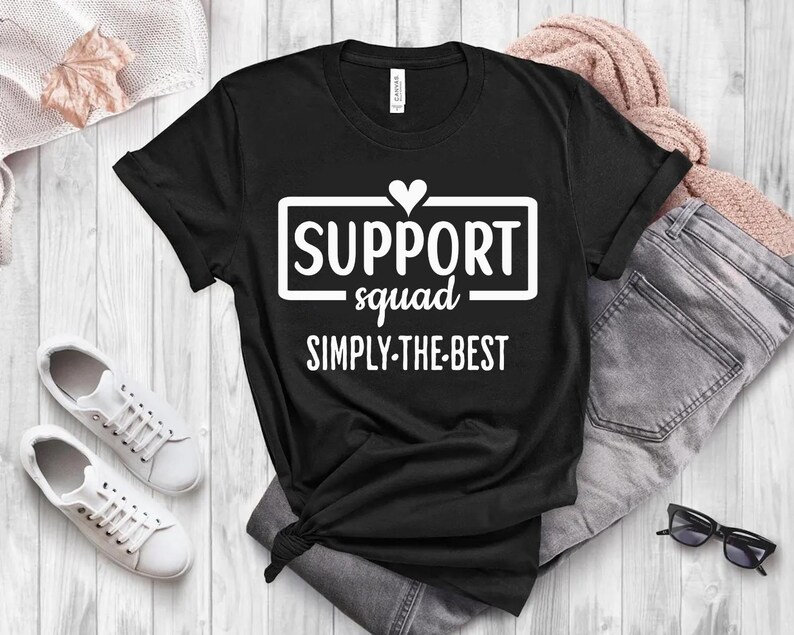 May include: Black t-shirt with the text "SUPPORT squad SIMPLY-THE-BEST" in a white rectangular box with a heart above the word "SUPPORT". The shirt is styled with a pair of white sneakers, blue jeans, and a pink sweater.