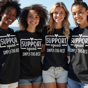 May include: Four women wearing black t-shirts with the text "SUPPORT squad SIMPLY-THE-BEST" in white. The shirts have a heart above the word "support". The women are smiling and standing in front of a blurred background.