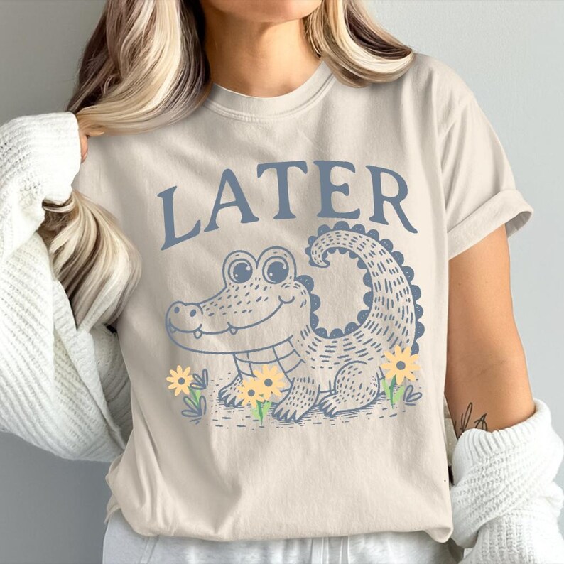 May include: A beige t-shirt featuring a blue alligator graphic and the word "LATER" printed in blue above the alligator.
