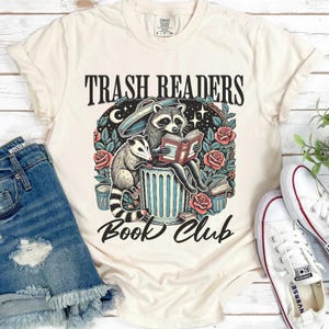 May include: A beige t-shirt with a graphic of a raccoon and opossum reading a book in a trash can. The text "Trash Readers Book Club" is printed on the shirt.