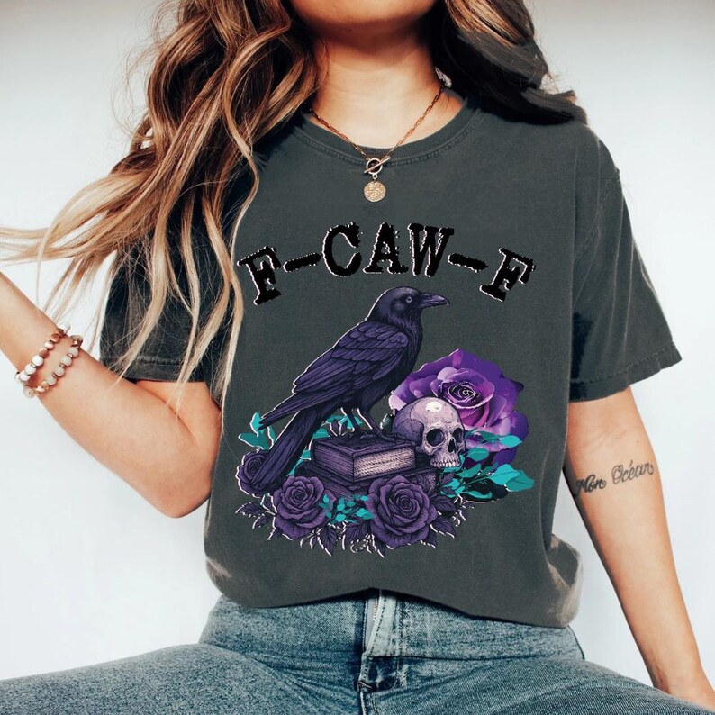 May include: Dark grey t-shirt featuring a graphic of a black crow perched above a skull, books, and purple roses. The text "F-CAW-F" is arched above the crow. The design has a gothic or alternative aesthetic.