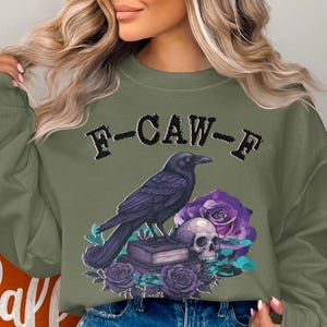 May include: Olive green sweatshirt with a graphic featuring a black crow perched on a stack of books, a skull, and purple roses. The text "F-CAW-F" is above the crow. A fall-themed design.