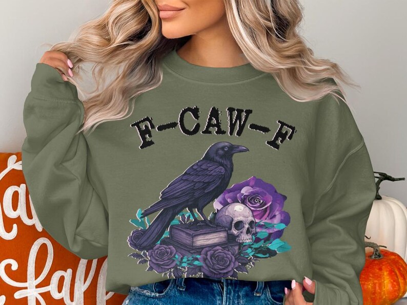 May include: Olive green sweatshirt with a graphic featuring a black crow perched on a stack of books, a skull, and purple roses. The text "F-CAW-F" is above the crow. A fall-themed design.
