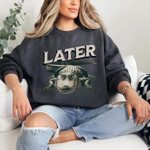 May include: A dark gray sweatshirt with a graphic of an alligator and the word "LATER" printed in white.