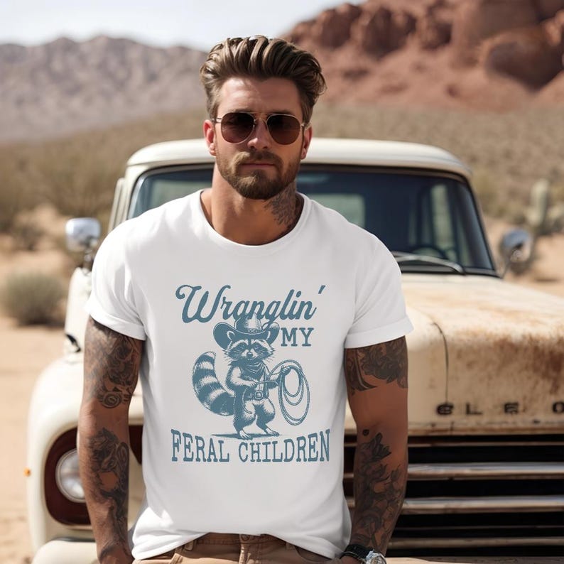 May include: White t-shirt with a blue graphic of a raccoon wearing a cowboy hat and holding a lasso. The text "Wranglin' My Feral Children" is printed below the graphic.