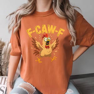 May include: Orange t-shirt featuring a cartoon chicken with the text "F-CAW-F". The chicken is depicted with its wings spread and mouth open in a surprised expression. The design is in shades of brown, yellow, and red.