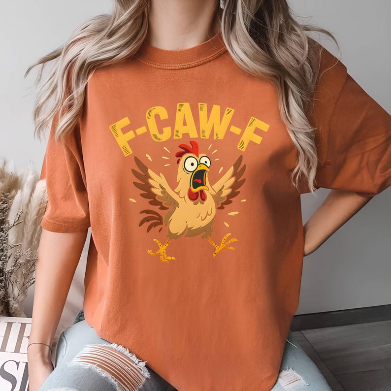 May include: Orange t-shirt featuring a cartoon chicken with the text "F-CAW-F". The chicken is depicted with its wings spread and mouth open in a surprised expression. The design is in shades of brown, yellow, and red.