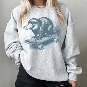 May include: A gray sweatshirt with a graphic of a platypus riding a skateboard. The platypus is blue and the skateboard is blue with black wheels.