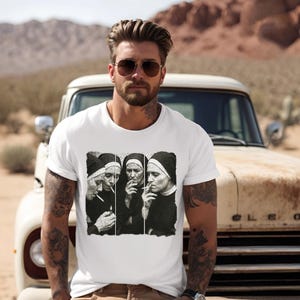 May include: White crew neck t-shirt featuring a black and white graphic of four nuns smoking cigarettes. The graphic is centered on the shirt. The background shows a man wearing sunglasses and a vintage truck.