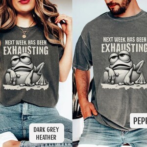 May include: A dark grey heather t-shirt with the text "Next week has been exhausting" and a cartoon frog with a tired expression. The shirt is available in a pepper color.